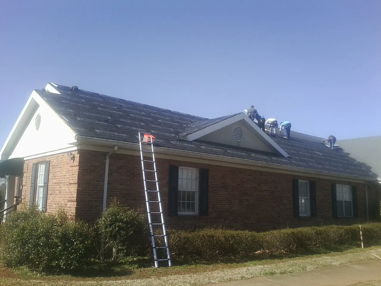 Crew tearing shingles off a brick ranch roof with a ladder set — Emergency Roof Repair work by our crew in Empire