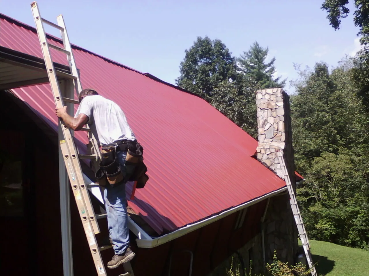 Roofer on a ladder beside a red metal roof and stone chimney — Soffit & Fascia Repair by Roofing Brothers in Empire